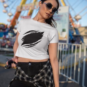 May include: A white cropped t-shirt with a black graphic of a ripped or torn design. The shirt is worn by a person with black shorts and a plaid shirt tied around their waist.