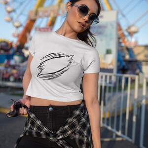 May include: A white crop top with a black graphic design of a jagged circle with a blank space in the center. The model is wearing black shorts and a plaid shirt tied around her waist. She is wearing sunglasses and standing in front of a Ferris wheel.