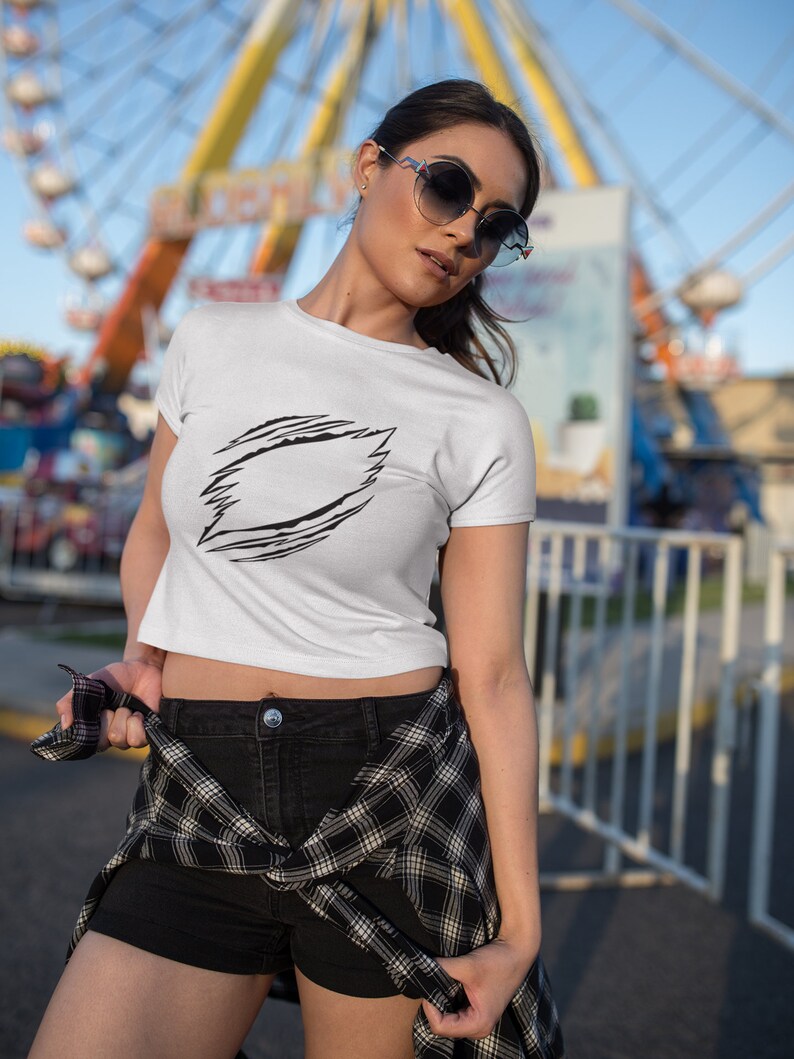 May include: A white crop top with a black graphic design of a jagged circle with a blank space in the center. The model is wearing black shorts and a plaid shirt tied around her waist. She is wearing sunglasses and standing in front of a Ferris wheel.