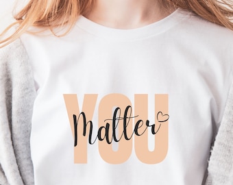 以下が含まれることがあります： 白いTシャツに、黒い文字で「YOU matter」と書いてあり、小さなハートのマークが付いています。テキストはピーチ色のボックスに入っています。