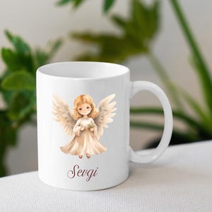 May include: White ceramic mug with a watercolour illustration of an angel with blonde hair, wings, and a cream-coloured dress. The word "Sevgi" is written in a cursive font at the bottom. The mug has a curved handle.