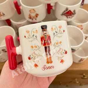 May include: White ceramic mug with a red handle, featuring a nutcracker illustration in red, black, and gold. The mug has floral accents and the name "Soner" in script. Other mugs with similar designs are in the background.