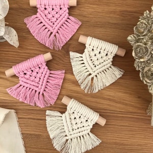 Puede incluir: Cuatro posavasos de macramé en rosa y crema con detalles de clavijas de madera. Los posavasos tienen un diseño geométrico con detalles de flecos. Están dispuestos sobre una superficie de madera, mostrando una estética artesanal.