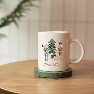 May include: White ceramic mug with a Christmas tree design, two nutcracker figures, and the text "Mutlu Yillar" in red. The mug is on a green marble coaster. The handle is white and the background is blurred.