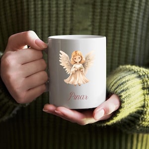 May include: A white ceramic mug with a cartoon angel design. The angel has blonde hair, wings, and a cream dress. The name "Pinar" is written in cursive. The mug is held by a person wearing a green jumper.