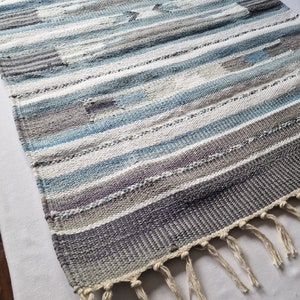 May include: A handwoven rug with a striped pattern in shades of blue, grey, and white. The rug has a fringed edge and a textured appearance, suitable for home decor.