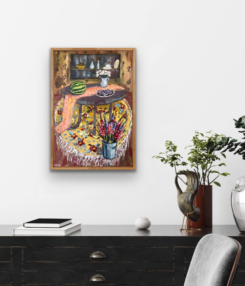 May include: A framed painting depicts a still life scene. A round table is covered with a yellow floral tablecloth and a pink scarf. A watermelon, blueberries, and a vase of flowers are on the table. A bucket of flowers sits below the table.