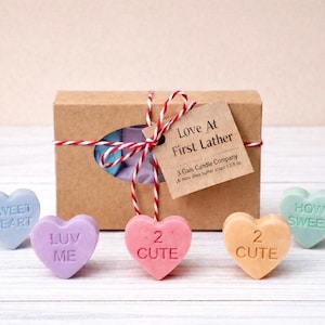 May include: A gift set featuring a brown box tied with red and white twine, and a tag that reads "Love At First Lather." Five heart-shaped soaps in pastel colors are arranged in front of the box, each with a different message.