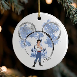 May include: A white ceramic Christmas ornament featuring a cartoon illustration of Prince Eric from The Little Mermaid. The design includes blue watercolor accents, a castle, and the name "Eric". The ornament hangs from a gold string.