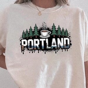 May include: Cream-colored t-shirt featuring a graphic of a coffee cup, pine trees, and the word "PORTLAND" in black, blue, and white. The design has a dripping effect. The shirt is short-sleeved.