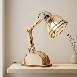 May include: A wooden desk lamp with a light bulb encased in a cage-like shade. The lamp has a natural wood finish and an adjustable arm. It sits on a wooden base and is plugged into an electrical outlet. The lamp is on a wooden shelf.