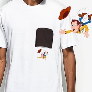 May include: White t-shirt featuring a cartoon illustration of a character falling out of a pocket. The character is wearing a cowboy hat, yellow shirt, blue jeans, and brown boots. The pocket is dark brown.