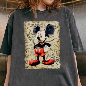 Może przedstawiać: Ciemnoszara koszulka z nadrukiem Mickey Mouse w stylu vintage. Myszka Miki jest przedstawiona w swoim klasycznym stroju z czerwonymi spodenkami i butami, czarną koszulą i białymi rękawiczkami. Tło zawiera tekst w stylu graffiti.