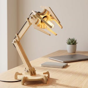 Könnte beinhalten: Eine Holz-Schreibtischlampe mit einzigartigem Design. Die Lampe hat eine Glühbirne, die von einem Holzrahmen mit Gelenken gehalten wird. Der Sockel und der Arm sind aus hellem Holz gefertigt. Ein Laptop und eine Sukkulentenpflanze befinden sich im Hintergrund.