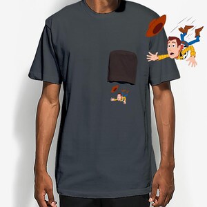 May include: Dark gray t-shirt featuring a cartoon illustration of a cowboy character falling from a pocket. The cowboy is wearing a yellow shirt, blue jeans, and a brown hat. The pocket is brown.