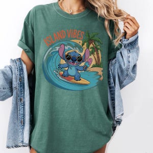 Puede incluir: Camiseta verde con el texto "ISLAND VIBES" sobre un personaje de dibujos animados surfeando una ola. El personaje azul lleva gafas de sol y sostiene un helado. Un cocotero y una escena de playa est&aacute;n en el fondo.