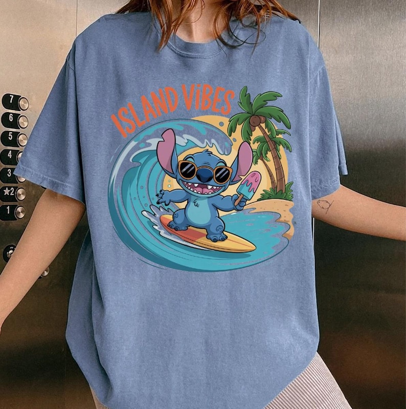 Puede incluir: Camiseta azul claro con un personaje de dibujos animados surfeando una ola, sosteniendo un helado. El gr&aacute;fico incluye el texto "ISLAND VIBES" en naranja. Palmeras y una puesta de sol en el fondo.