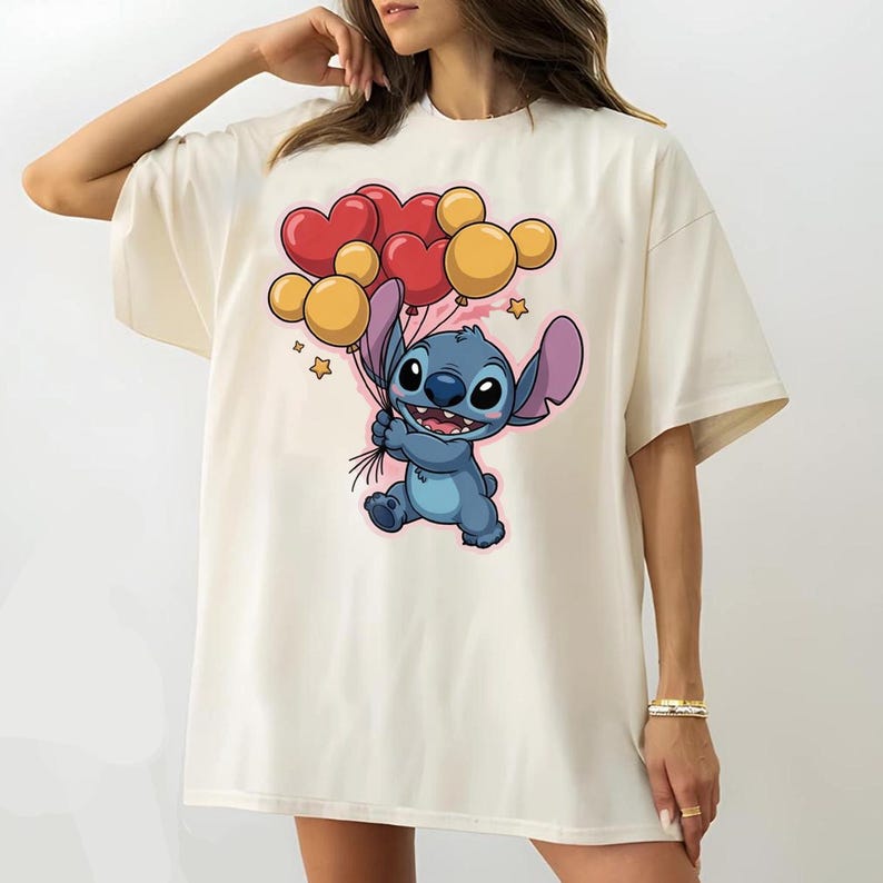 May include: Cream-colored t-shirt featuring a cartoon character holding heart and round balloons. The character is blue with large ears and a friendly expression. The balloons are red and yellow, with small star accents.