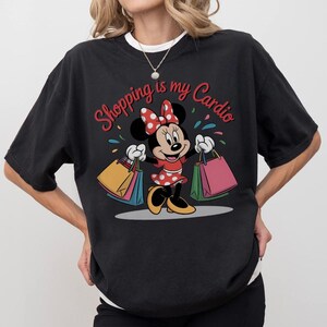 Puede incluir: Camiseta negra con Minnie Mouse sosteniendo bolsas de compras. Minnie lleva un vestido rojo con lunares blancos y un lazo a juego. El texto "Shopping is my Cardio" est&aacute; escrito en rojo sobre el personaje.