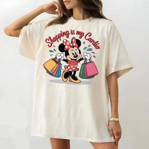 Puede incluir: Camiseta color crema con una Minnie Mouse de dibujos animados sosteniendo bolsas de compras. Minnie Mouse lleva un vestido y un lazo de lunares rojos. El texto "Shopping is my Cardio" est&aacute; escrito sobre el personaje en rojo.