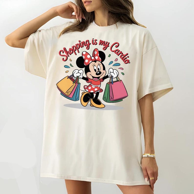 Puede incluir: Camiseta color crema con una Minnie Mouse de dibujos animados sosteniendo bolsas de compras. Minnie Mouse lleva un vestido y un lazo de lunares rojos. El texto "Shopping is my Cardio" est&aacute; escrito sobre el personaje en rojo.