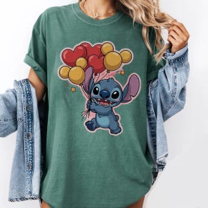 May include: Green t-shirt featuring a cartoon illustration of Stitch holding heart and Mickey Mouse-shaped balloons. The balloons are red and yellow. The shirt has a crew neck and short sleeves.