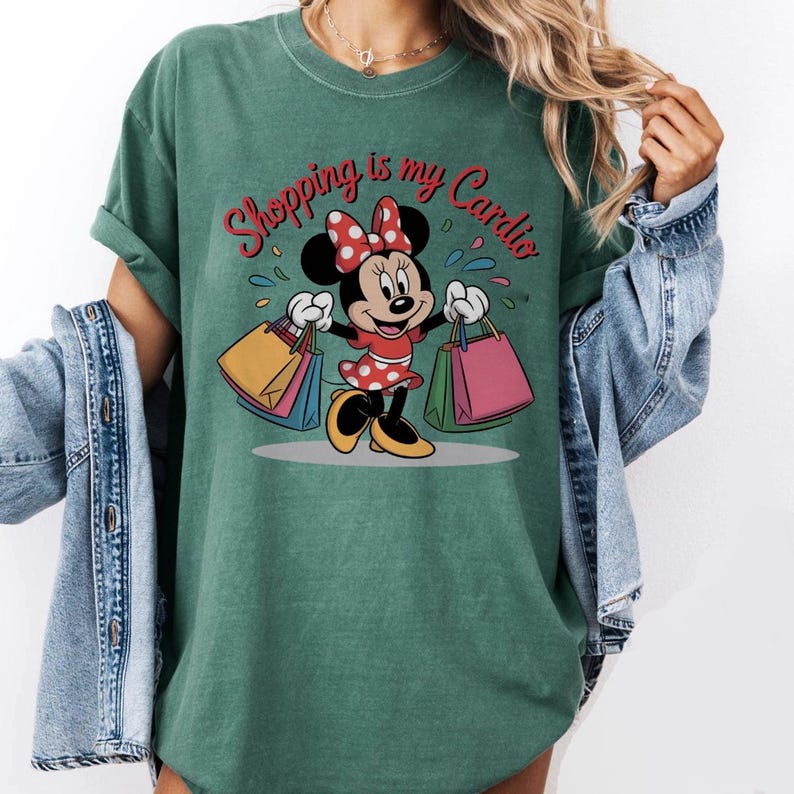 Puede incluir: Camiseta verde con un dise&ntilde;o de Minnie Mouse sosteniendo bolsas de compras. Minnie Mouse lleva un vestido rojo con lunares blancos y un lazo a juego. El texto "Shopping is my Cardio" est&aacute; escrito encima del personaje.
