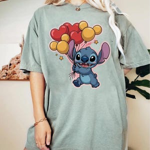 May include: A light green t-shirt featuring a cartoon character holding heart and round balloons. The character is blue with large ears and a friendly expression. The balloons are red and yellow.