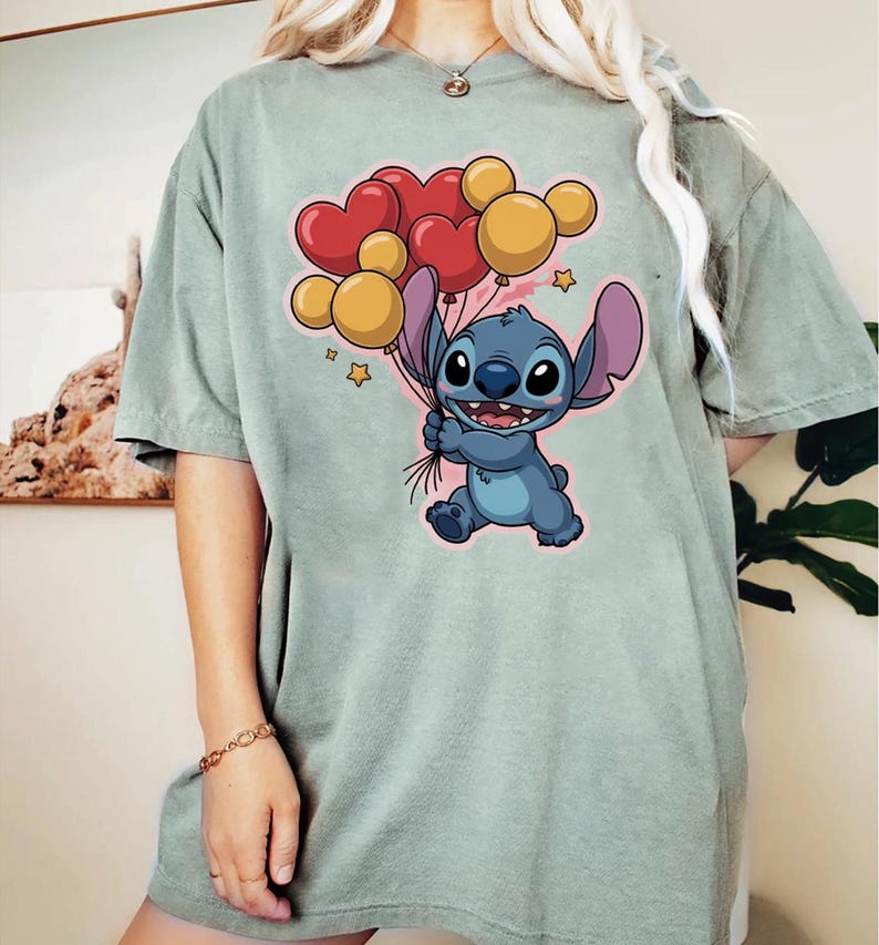 May include: A light green t-shirt featuring a cartoon character holding heart and round balloons. The character is blue with large ears and a friendly expression. The balloons are red and yellow.