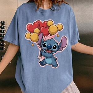 May include: A light blue t-shirt featuring a cartoon character holding heart and round balloons. The character is blue with large ears and a friendly expression. The balloons are red and yellow.