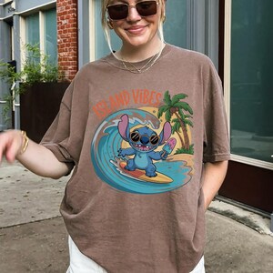 Puede incluir: Camiseta marr&oacute;n con un dise&ntilde;o de Stitch surfeando una ola, con el texto "Island Vibes". El dise&ntilde;o incluye una palmera y Stitch sosteniendo un helado. La camiseta tiene un corte holgado.