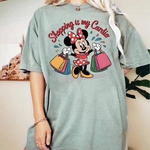 Puede incluir: Camiseta verde salvia con Minnie Mouse sosteniendo bolsas de compras. El gr&aacute;fico incluye el texto "Shopping is my Cardio" en rojo y blanco. Minnie Mouse lleva un vestido rojo con lunares blancos y un lazo a juego.