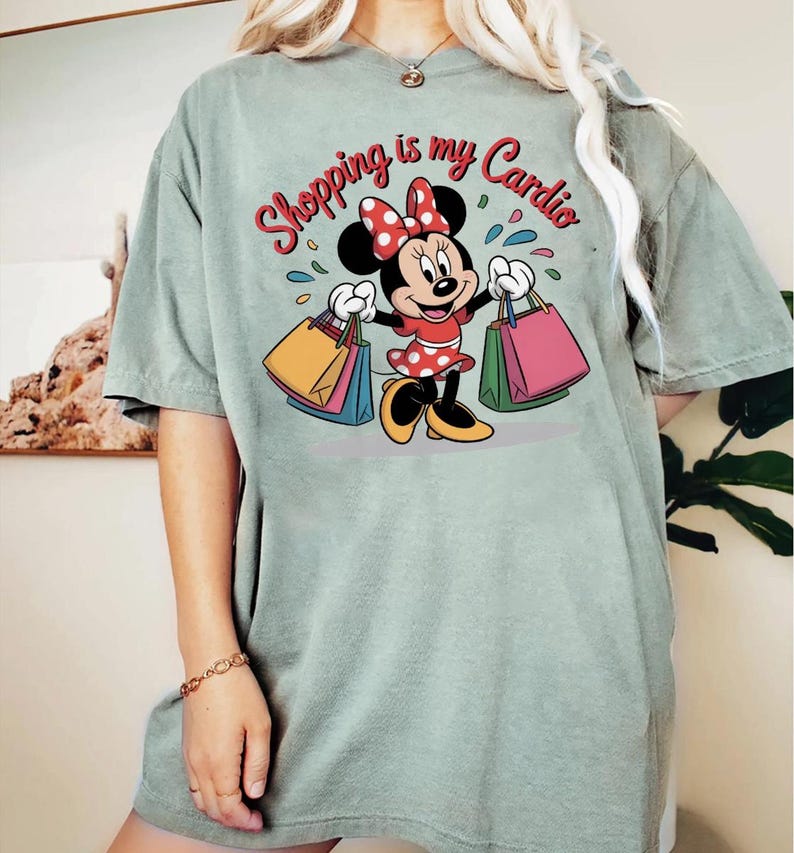 Puede incluir: Camiseta verde salvia con Minnie Mouse sosteniendo bolsas de compras. El gr&aacute;fico incluye el texto "Shopping is my Cardio" en rojo y blanco. Minnie Mouse lleva un vestido rojo con lunares blancos y un lazo a juego.
