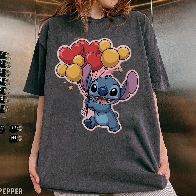 May include: Dark gray t-shirt featuring a cartoon Stitch character holding heart and round balloons. The balloons are red and yellow. The design includes small yellow stars. The shirt is a casual, oversized style.