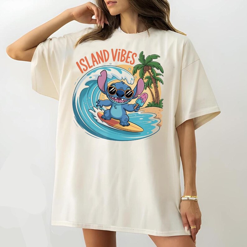 Puede incluir: Camiseta color crema con un personaje de dibujos animados surfeando una ola, sosteniendo un helado. El gr&aacute;fico incluye el texto "ISLAND VIBES" en naranja. Tambi&eacute;n se presentan palmeras y una escena de playa.
