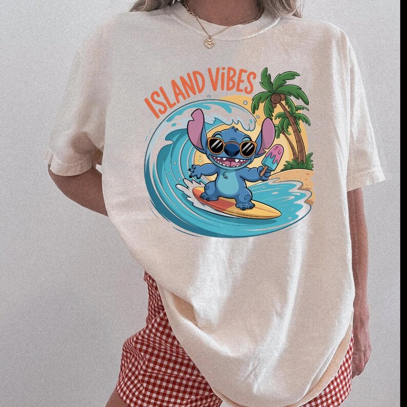 Puede incluir: Camiseta color crema con un personaje de dibujos animados surfeando una ola y sosteniendo un helado. El gr&aacute;fico incluye el texto "ISLAND VIBES" en naranja. Una palmera y una escena de playa completan el dise&ntilde;o.
