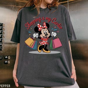 Puede incluir: Camiseta gris oscuro con Minnie Mouse sosteniendo bolsas de compras. Minnie Mouse lleva un vestido rojo con lunares blancos y un lazo a juego. El texto "Shopping is my Cardio" est&aacute; encima de Minnie.