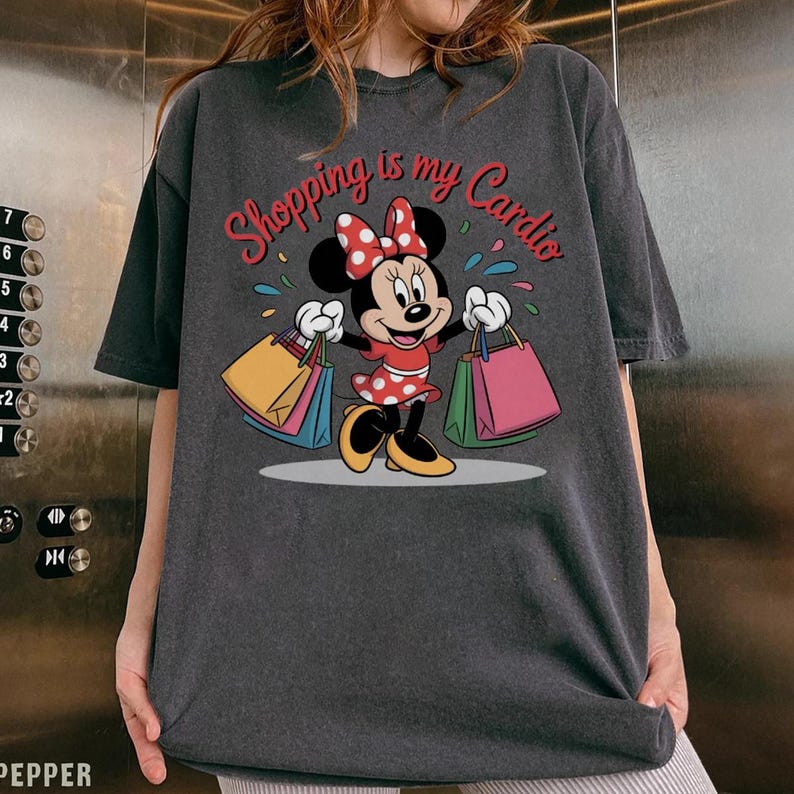 Puede incluir: Camiseta gris oscuro con Minnie Mouse sosteniendo bolsas de compras. Minnie Mouse lleva un vestido rojo con lunares blancos y un lazo a juego. El texto "Shopping is my Cardio" est&aacute; encima de Minnie.