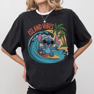 Puede incluir: Camiseta negra con un gr&aacute;fico de un personaje de dibujos animados azul surfeando una ola, sosteniendo un helado y usando gafas de sol. Las palabras "Island Vibes" est&aacute;n impresas encima del gr&aacute;fico. Una palmera y una playa est&aacute;n en el fondo.