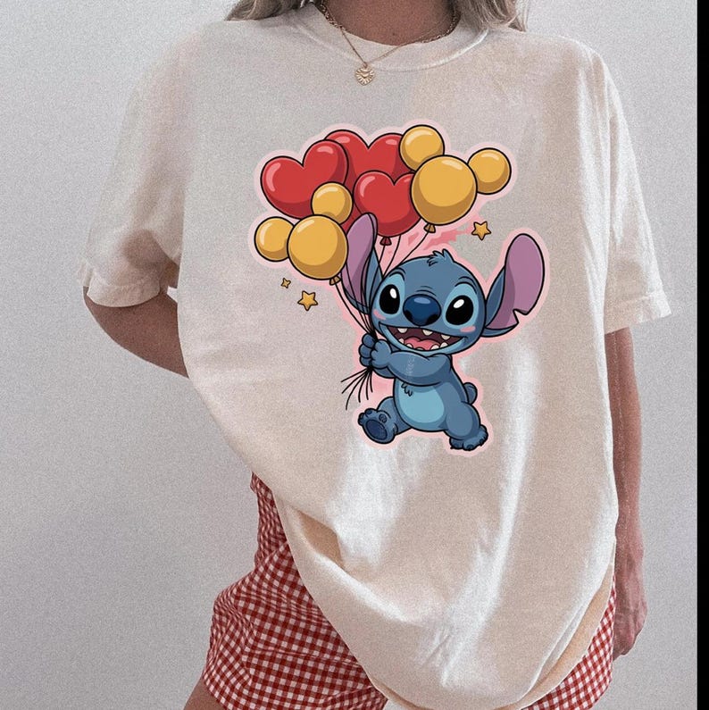 May include: Cream-colored t-shirt featuring a cartoon character holding heart and round balloons. The character is blue with large ears and a wide smile. The balloons are red and yellow. Small stars are also included in the design.