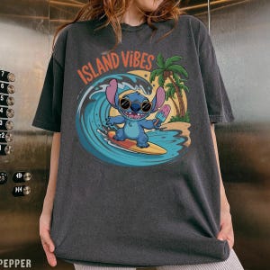 Puede incluir: Camiseta gris oscuro con un personaje de dibujos animados surfeando una ola, sosteniendo un helado. El gr&aacute;fico incluye el texto "ISLAND VIBES" en naranja, una palmera y una escena de playa. El personaje es azul y lleva gafas de sol.
