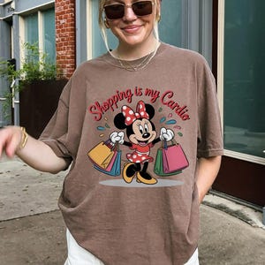 Puede incluir: Camiseta marr&oacute;n con un dise&ntilde;o de Minnie Mouse sosteniendo bolsas de compras. El texto "Shopping is my Cardio" est&aacute; impreso encima del personaje. La camiseta tiene mangas cortas y un corte holgado.