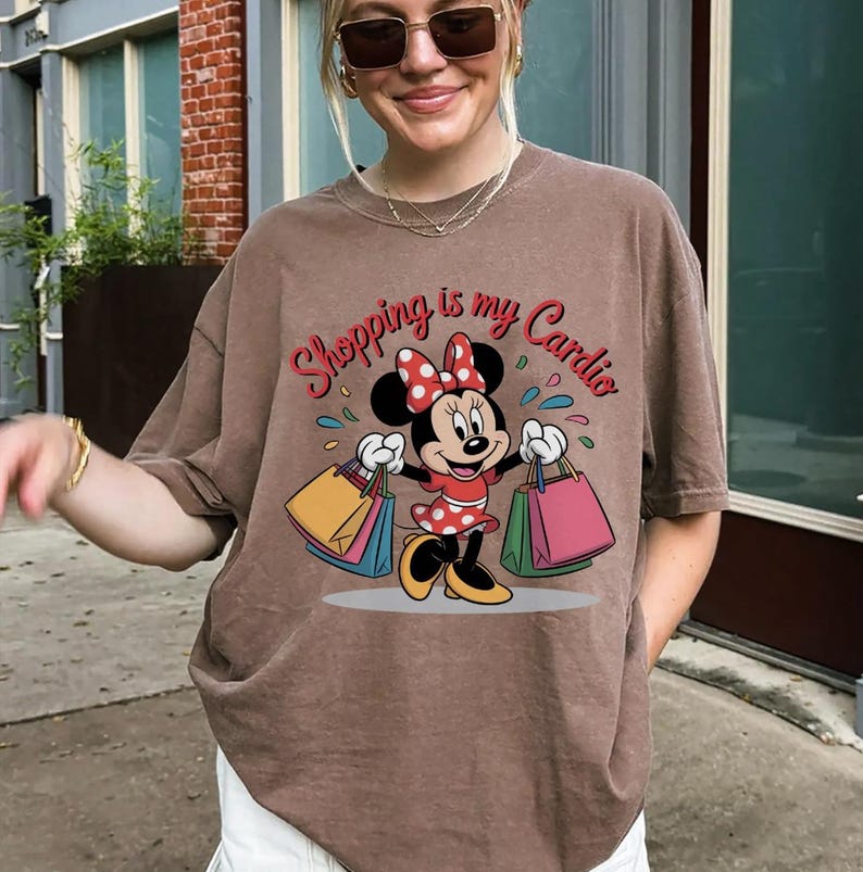 Puede incluir: Camiseta marr&oacute;n con un dise&ntilde;o de Minnie Mouse sosteniendo bolsas de compras. El texto "Shopping is my Cardio" est&aacute; impreso encima del personaje. La camiseta tiene mangas cortas y un corte holgado.