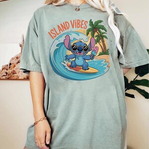 Puede incluir: Camiseta verde salvia con un personaje de dibujos animados azul surfeando una ola, sosteniendo un helado. El texto "ISLAND VIBES" est&aacute; arqueado sobre el personaje. Un dise&ntilde;o de palmera y escena de playa.