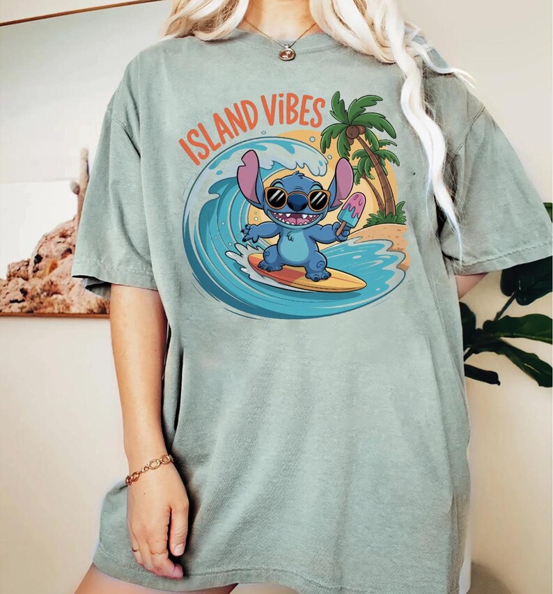 Puede incluir: Camiseta verde salvia con un personaje de dibujos animados azul surfeando una ola, sosteniendo un helado. El texto "ISLAND VIBES" est&aacute; arqueado sobre el personaje. Un dise&ntilde;o de palmera y escena de playa.