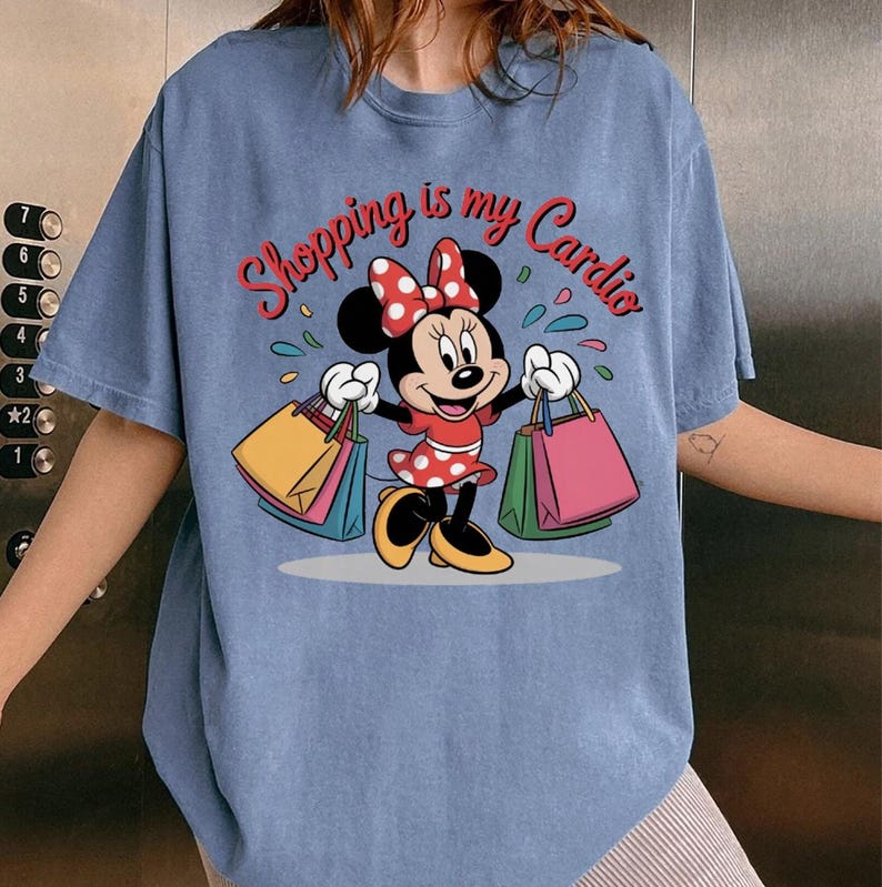 Puede incluir: Camiseta azul claro con Minnie Mouse sosteniendo bolsas de compras. El texto "Shopping is my Cardio" est&aacute; en rojo. Minnie Mouse lleva un vestido rojo con lunares blancos y un lazo a juego. Estilo casual.