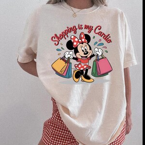 Puede incluir: Camiseta color crema con un dibujo animado de Minnie Mouse sosteniendo bolsas de compras. El texto "Shopping is my Cardio" est&aacute; escrito encima del personaje en rojo y rosa. La camiseta se combina con unos pantalones cortos a cuadros rojos y blancos.
