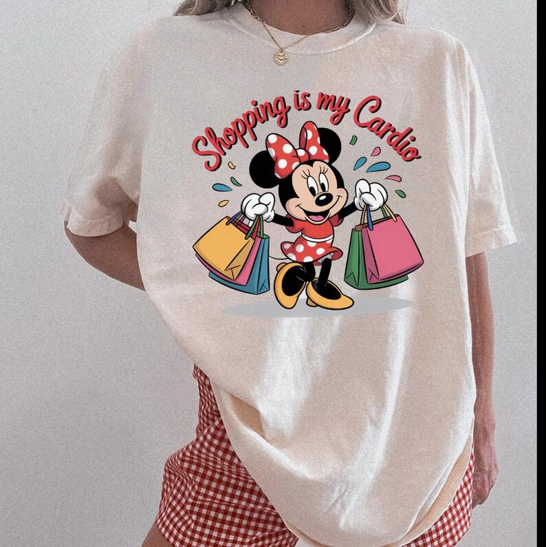 Puede incluir: Camiseta color crema con un dibujo animado de Minnie Mouse sosteniendo bolsas de compras. El texto "Shopping is my Cardio" est&aacute; escrito encima del personaje en rojo y rosa. La camiseta se combina con unos pantalones cortos a cuadros rojos y blancos.