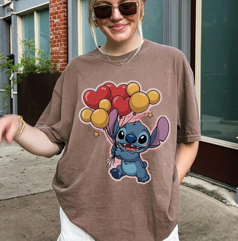 May include: Brown t-shirt featuring a cartoon character holding heart and round balloons. The character is blue with large ears and a friendly expression. The balloons are red and yellow, with small star accents.