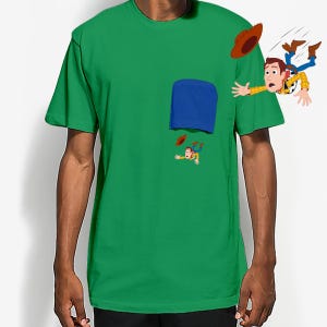 May include: Green t-shirt featuring a blue pocket design with a cartoon illustration of a character falling out of the pocket. The character is wearing a cowboy hat, a yellow shirt, and blue jeans.